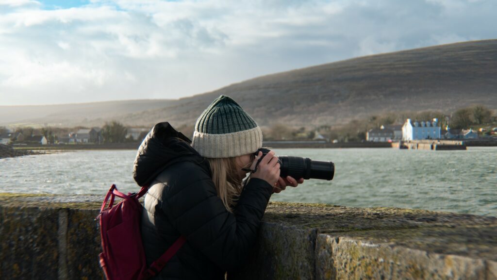 Blonde girl with hat looks through a camera to take a photo overlooking the water in Ireland.