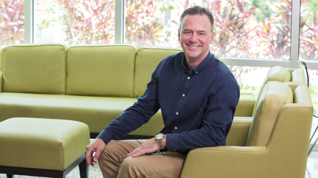 A smiling man poses for a picture while sitting on a green leather armchair.